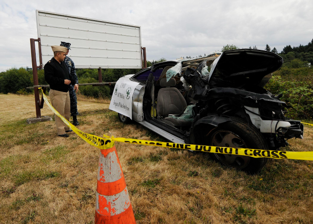 Ein Polizist in Uniform steht neben einem schwer beschädigten Auto am Straßenrand, mit einem Verkehrskegel und einem Band an einem Pfahl in der Nähe, sowie Bäumen und einem bewölkten Himmel im Hintergrund.