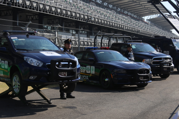 Eine Gruppe von Polizeiwagen steht vor einer Rennstrecke mit uniformierten Personen auf der Straße, im Hintergrund ein Stadion und Maschendrahtzaun.