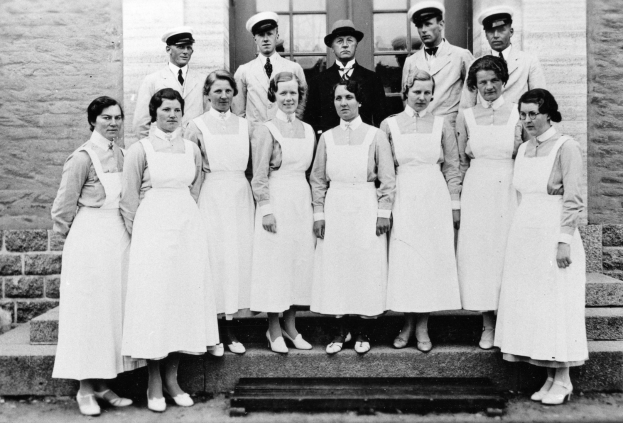 Eine Gruppe von Schwestern, die auf den Stufen eines Gebäudes stehen, mit einer Wand und Fenstern im Hintergrund, dargestellt in Schwarz-Weiß.